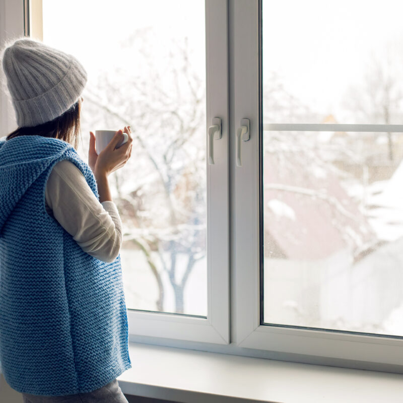 A woman in a soft blue knit sweater holding a warm drink, gazing out a window at a peaceful snowy scene.